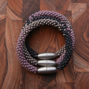 Puede incluir: Un brazalete de cuentas multicadena con cierre magnético. El brazalete está hecho de pequeñas cuentas redondas en tonos de morado, gris y negro.