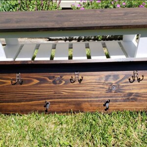 May include: A wooden entryway bench with a dark brown top and white slatted base. Below, a matching wooden coat rack with six metal hooks is displayed. The items are set on a grassy surface.