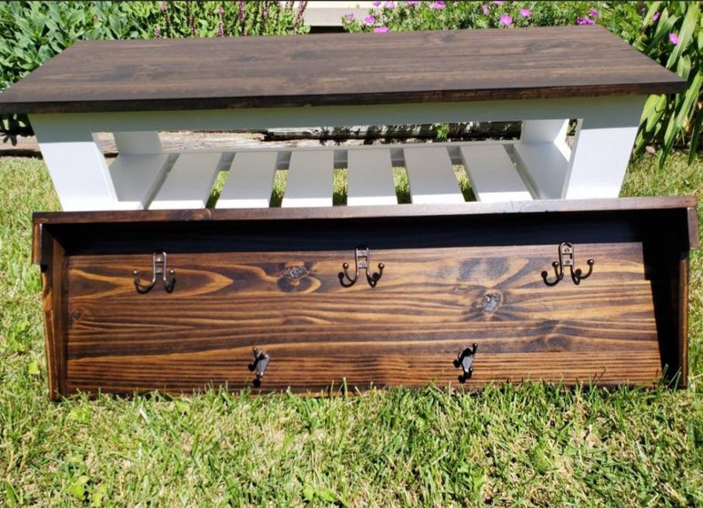 May include: A wooden entryway bench with a dark brown top and white slatted base. Below, a matching wooden coat rack with six metal hooks is displayed. The items are set on a grassy surface.