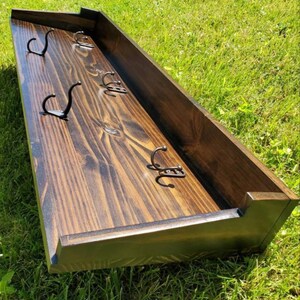 May include: A dark stained wooden wall-mounted coat rack with six metal hooks. The rack has a rectangular shape with a raised border and is set against a green grassy background. The hooks are evenly spaced.