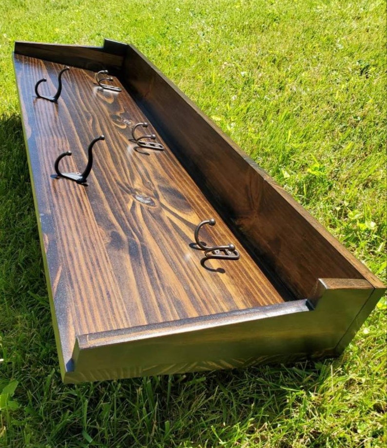 May include: A dark stained wooden wall-mounted coat rack with six metal hooks. The rack has a rectangular shape with a raised border and is set against a green grassy background. The hooks are evenly spaced.
