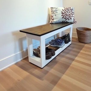 May include: A wooden shoe bench with a white base and a dark brown top. The bench has a lower shelf for shoe storage. A decorative pillow rests on the bench. Shoes are visible on the shelf.