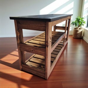 Op de afbeelding: Houten keukeneiland met een donker gebeitst bovenblad en twee onderste planken. Het eiland is gemaakt van massief hout met een natuurlijke afwerking, waarbij de houtnerf zichtbaar is. Het ontwerp is rustiek en functioneel, geschikt voor een keuken of eetkamer. Ongeveer 120 cm lang.