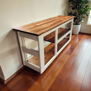 May include: A wooden console table with a white frame and two shelves. The top and shelves are made of stained wood. The table is positioned against a beige wall and sits on a hardwood floor.
