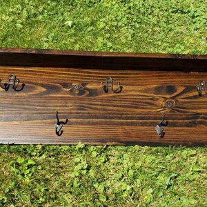 May include: A dark stained wooden wall shelf with six metal hooks. The shelf has a rectangular shape with a raised back and is set against a background of green grass. The hooks are evenly spaced.