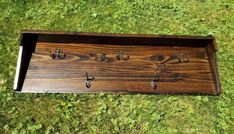 May include: A dark stained wooden wall shelf with six metal hooks. The shelf has a rectangular shape with a raised back and is set against a background of green grass. The hooks are evenly spaced.