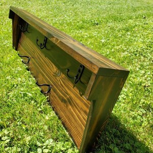 May include: A wooden wall-mounted coat rack with a dark stain finish. It features a sloped top and several black metal hooks for hanging items. The rack is set against a backdrop of green grass.