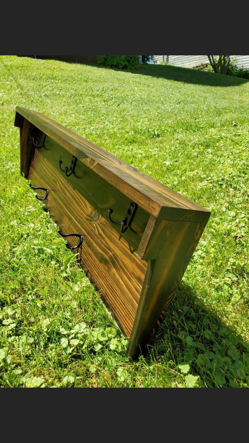 May include: A wooden wall-mounted coat rack with a dark stain finish. It features a sloped top and several black metal hooks for hanging items. The rack is set against a backdrop of green grass.