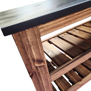Op de afbeelding: Houten bank met een donkergrijze bovenkant en een onderste plank van houten latten. De bank heeft een natuurlijke houten afwerking met zichtbare nerven en knopen. De poten zijn schuin voor stabiliteit.