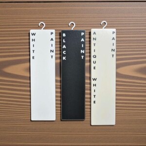 May include: Three rectangular paint color samples labeled "WHITE PAINT", "BLACK PAINT", and "ANTIQUE WHITE PAINT" hang against a wood grain background. Each sample has a metal hook at the top.