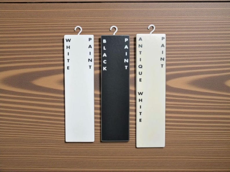 May include: Three rectangular paint color samples labeled "WHITE PAINT", "BLACK PAINT", and "ANTIQUE WHITE PAINT" hang against a wood grain background. Each sample has a metal hook at the top.