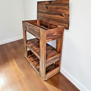 May include: A wooden shoe rack with a dark stained finish. The rack has two shelves and a top that can be lifted. The wood grain is visible. The rack is placed against a white wall and sits on a hardwood floor.