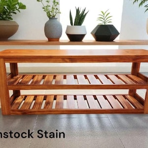 May include: A wooden shoe rack with two slatted shelves and a solid top. The rack is stained in a warm gunstock color. The text "Gunstock Stain" is at the bottom of the image. The rack is in front of a shelf with plants.