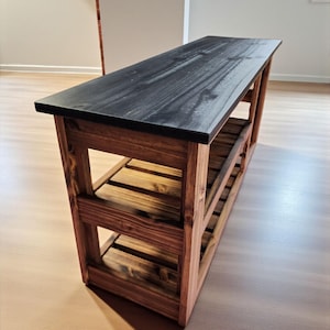Op de afbeelding: Houten bank met een zwart blad en een natuurlijke houten basis. De bank heeft twee onderste planken en is opgebouwd met een lattenontwerp. Afmetingen: ongeveer 122 cm lang, 36 cm breed en 46 cm hoog.