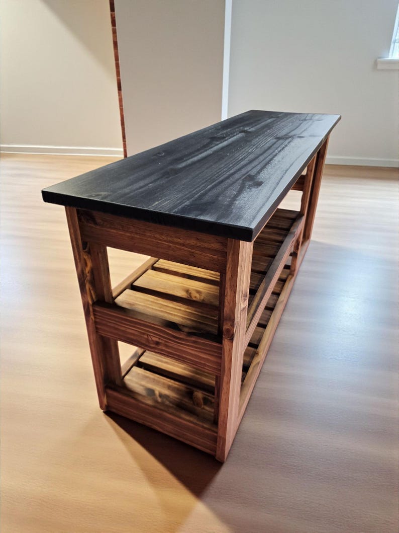 Op de afbeelding: Houten bank met een zwart blad en een natuurlijke houten basis. De bank heeft twee onderste planken en is opgebouwd met een lattenontwerp. Afmetingen: ongeveer 122 cm lang, 36 cm breed en 46 cm hoog.