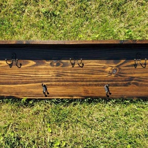May include: A dark stained wooden wall shelf with four double hooks. The shelf has a rectangular shape with a raised back and is set against a background of green grass.