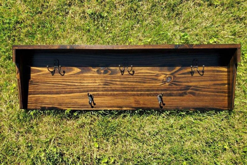 May include: A dark stained wooden wall shelf with four double hooks. The shelf has a rectangular shape with a raised back and is set against a background of green grass.