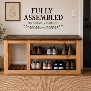 May include: A wooden shoe bench with a dark brown top and a natural light wood frame, featuring two open shelves filled with various pairs of sneakers and boots. The wall above displays the text FULLY ASSEMBLED NO ASSEMBLY REQUIRED.