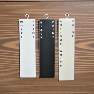 Op de afbeelding: Drie rechthoekige verfkleurstalen met de labels "WHITE PAINT", "BLACK PAINT" en "ANTIQUE WHITE PAINT" hangen tegen een achtergrond van houtnerf. Elk staal heeft een metalen haak aan de bovenkant.