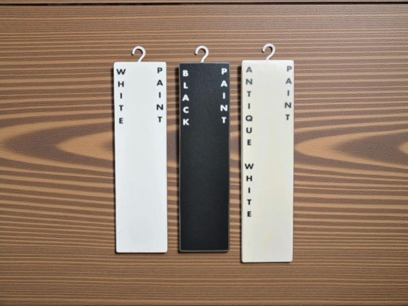 Op de afbeelding: Drie rechthoekige verfkleurstalen met de labels "WHITE PAINT", "BLACK PAINT" en "ANTIQUE WHITE PAINT" hangen tegen een achtergrond van houtnerf. Elk staal heeft een metalen haak aan de bovenkant.