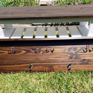 May include: A wooden bench with a dark brown top and white slatted shelf. Below, a dark brown wooden coat rack with six metal hooks is visible. The bench is approximately 48 inches long.