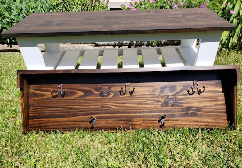 May include: A wooden bench with a dark brown top and white slatted shelf. Below, a dark brown wooden coat rack with six metal hooks is visible. The bench is approximately 48 inches long.