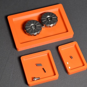 Puede incluir: Una vista aérea muestra una disposición de pequeñas piezas y componentes metálicos en bandejas de color naranja brillante. Dos objetos circulares con forma de engranaje se encuentran en una bandeja rectangular. Otras bandejas contienen piezas cilíndricas y en forma de tornillo más pequeñas. El fondo es gris oscuro.