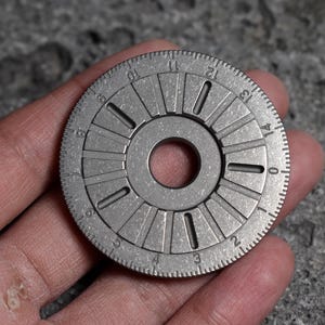 以下が含まれることがあります： 中央に穴と放射状のマークが付いた、銀色の円形金属オブジェクト。外側のエッジには番号付きの増分があり、表面はテクスチャ加工されています。オブジェクトは、灰色のテクスチャ加工された背景に対して、手で保持されています。
