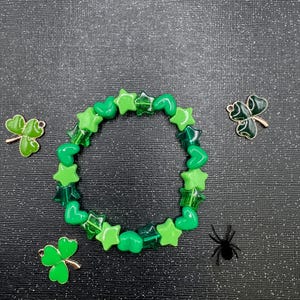 Può includere: Un braccialetto di perline verdi con perline a forma di stella e cuore. Tre ciondoli a forma di trifoglio e un ragno nero sono disposti attorno al braccialetto su una superficie scura e glitterata. Un accessorio festivo.