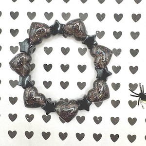 Puede incluir: Un brazalete con cuentas alternas en forma de estrella y corazón de color negro. Las cuentas de corazón son oscuras con purpurina, sobre un fondo blanco con patrones de corazones negros. Un accesorio de moda.