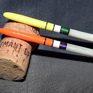 Könnte beinhalten: Zwei Angelposen mit weißen Körpern und grünen und violetten Spitzen. Die Posen liegen auf einem Korken.