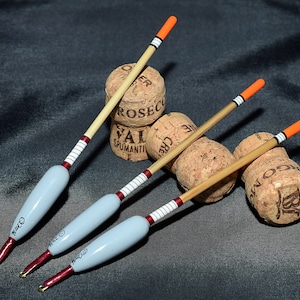 May include: Three wooden fishing floats with orange and white tips. The floats are resting on top of three champagne corks.