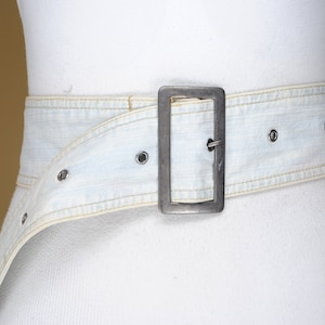 May include: Light blue denim belt with a large, rectangular silver-tone buckle. The belt has a wide design and is secured with metal grommets. The light wash denim is accented with yellow stitching.