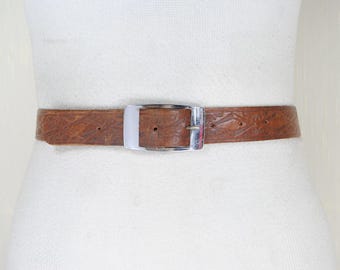 May include: A brown leather belt with a silver-colored rectangular buckle. The belt has a textured surface with a repeating geometric pattern. The belt is displayed on a white mannequin.