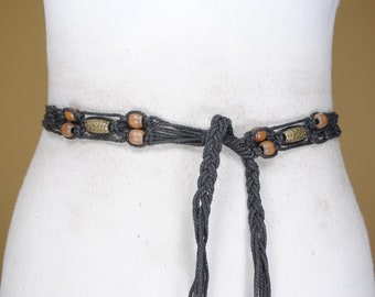 Puede incluir: Un cinturón de macramé gris oscuro con un diseño de cordón trenzado, intercalado con cuentas de madera redondas y cuentas metálicas texturizadas alargadas. El cinturón está atado en el centro con dos extremos de cordón trenzado largos.