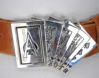 May include: A brown leather belt with a silver buckle featuring playing cards. The buckle design includes the Ace of Spades and other cards, with the letters K, Q, J, and 10 visible. The belt is secured with a silver buckle.
