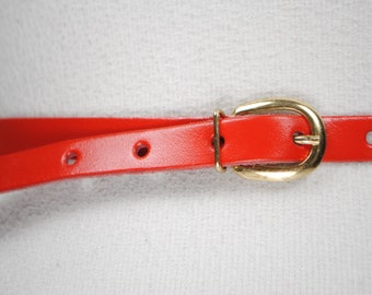 May include: A bright red belt with a gold-colored buckle. The belt is thin and has several holes for adjustment. The buckle is a classic oval shape. This belt is a fashion accessory.