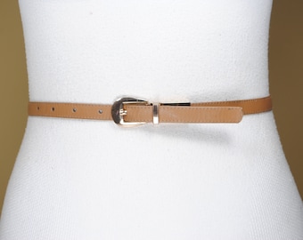 May include: A tan-colored belt with a gold-colored buckle is displayed on a white mannequin. The belt is thin and has a classic design. The buckle is rectangular with a rounded edge. The belt is approximately 1 inch wide.