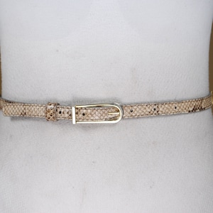 以下が含まれることがあります： ベージュと茶色のスネークスキン柄の細いベルト。バックルとループは金色です。白いマネキンにベルトが表示されています。