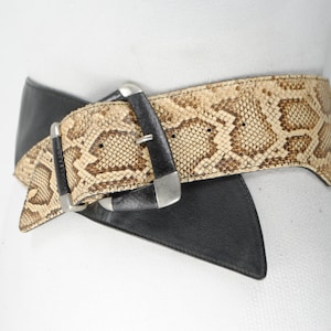 May include: A wide belt featuring a snakeskin pattern and black leather. The belt has two silver-toned buckles and a distinctive design. This fashion accessory is designed to be worn around the waist.