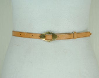 May include: A tan leather belt with a decorative gold-colored buckle. The belt is stitched with dark thread and has a classic design. The belt is shown on a mannequin.