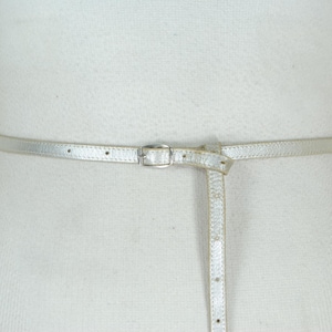 May include: A silver-colored belt with a small, rectangular buckle. The belt is thin and appears to be made of a metallic material. The belt is fastened around a white form, showcasing its design and fit. A stylish accessory.