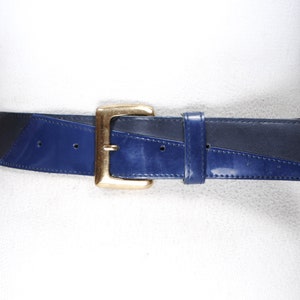 Puede incluir: Cinturón ancho y vintage con hebilla rectangular dorada. El cinturón es de cuero azul marino y azul oscuro, con un diseño geométrico. El cinturón se muestra en un maniquí blanco.