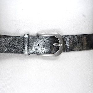 May include: A silver and black belt with a textured, reptile-like pattern. The belt has a silver-toned buckle and is displayed on a white mannequin. The belt is a fashion accessory.