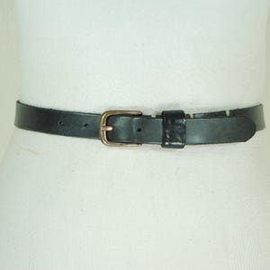 May include: A black leather belt with a brass buckle is shown. The belt is worn around a white form. The buckle is rectangular with a rounded edge. The belt has a single belt loop.
