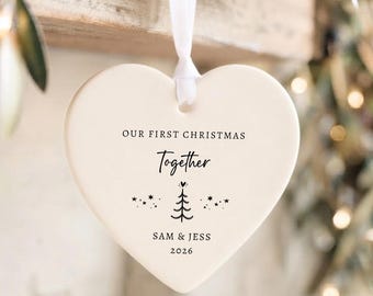 Pode incluir: Um ornamento de cerâmica branco em forma de coração pendurado por uma fita branca. O ornamento apresenta o texto preto "OUR FIRST CHRISTMAS Together" acima de um gráfico de árvore e estrelas, com os nomes "SAM & JESS" e o ano "2026" abaixo.