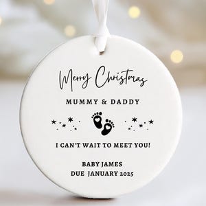 May include: White ceramic Christmas bauble with black text that reads "Merry Christmas Mummy & Daddy" and "I can't wait to meet you!" with a pair of footprints and stars. The bauble also includes the text "Baby James Due January 2025".