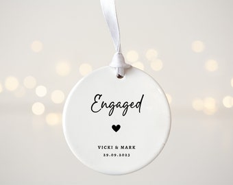 May include: White ceramic ornament with the word "Engaged" in black script, a small black heart, and the text "Vicki & Mark 29.09.2023" in black.