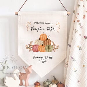 May include: A cream-coloured fabric wall banner hanging from a wooden dowel with a leather cord. The banner features a watercolour illustration of various pumpkins and the text "WELCOME TO OUR Pumpkin Patch" and "Mummy, Daddy & Isla" in black.