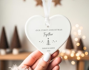 May include: A white heart-shaped ceramic ornament hanging from a white ribbon. The ornament features black text that reads OUR FIRST CHRISTMAS Together, a small line drawing of a Christmas tree with stars, and the names SARAH & JAMES with the year 2025.